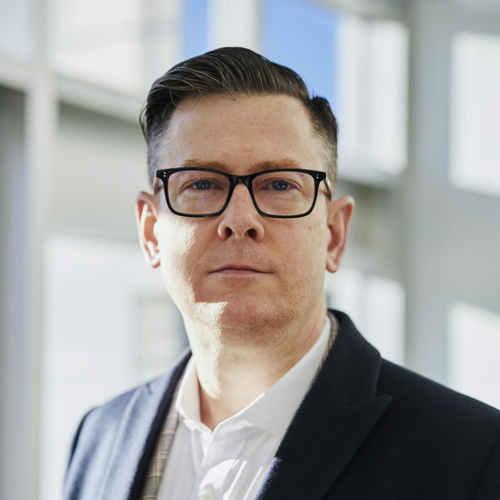Profile portrait of a confident business executive wearing eyeglasses in a sunlit office setting.
